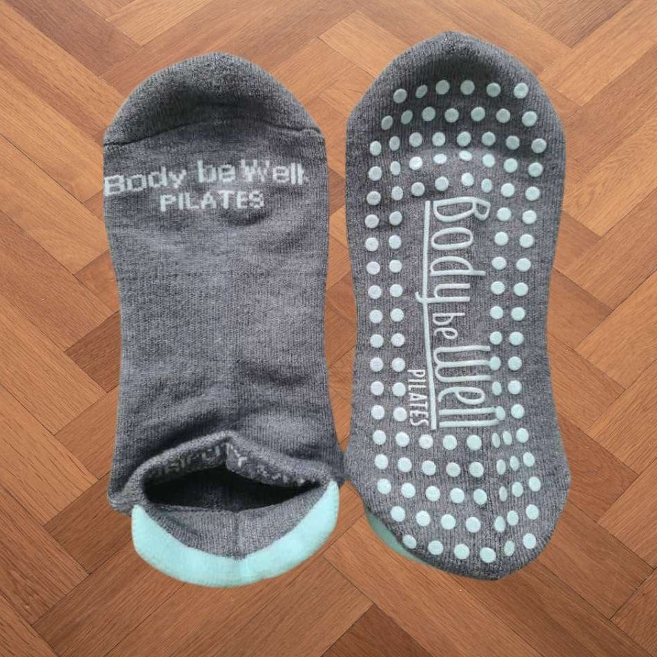 Gray pilates socks with textured sole on a wooden floor, branded 'Body be Well Pilates'.